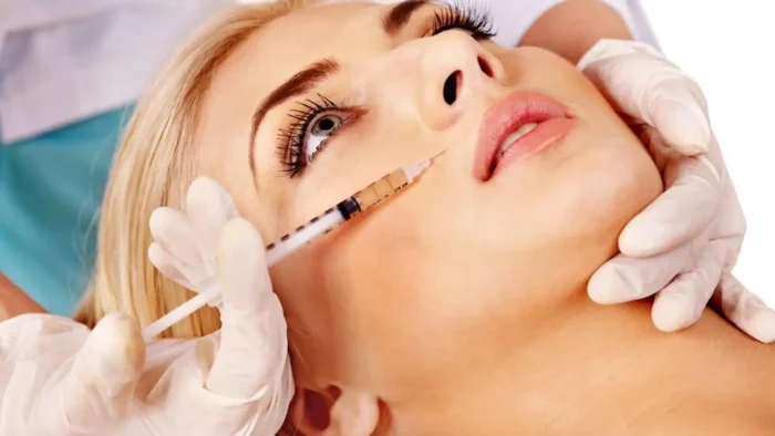 A close-up of a person receiving a cosmetic injection in the face. A healthcare professional wearing white gloves holds a syringe near the person's cheek. The person lies back with eyes open, in a clinical setting.