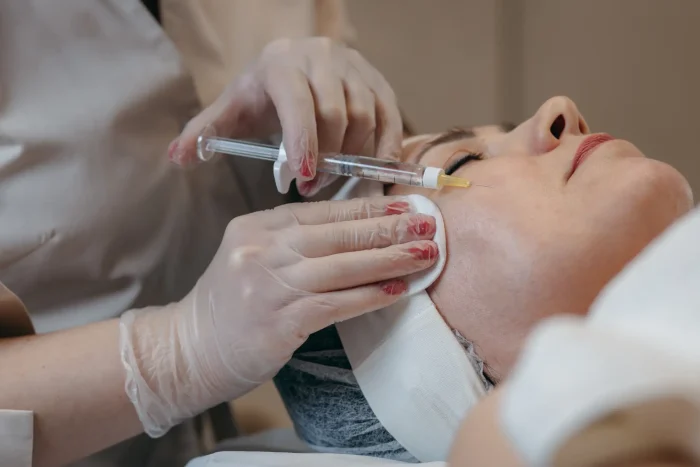A person receives a cosmetic facial injection in a clinical setting. A professional wearing gloves administers the treatment, with the patient lying on a bed, relaxed and with eyes closed.
