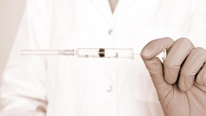 A person in a white coat holds a syringe with a measured dose of liquid. The image is in sepia tones, focusing on the gloved hand and the syringe.