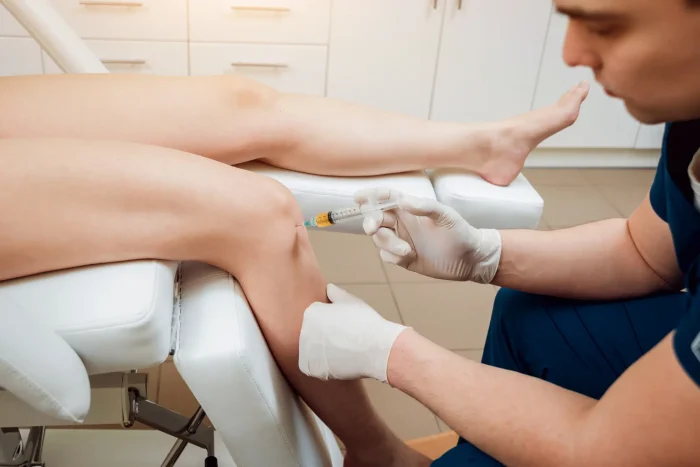 A person wearing gloves is administering an injection into a patient's knee while the patient is reclining on a medical chair in a clinic setting.