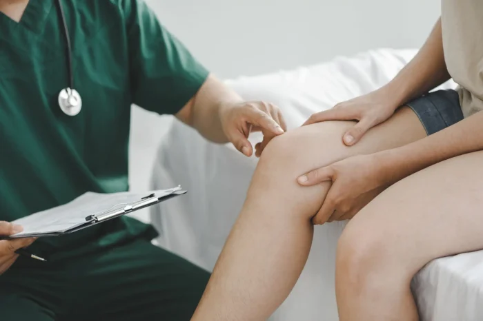 A healthcare professional examines a patient's knee. The patient is seated on an examination table, and the professional is wearing green scrubs with a stethoscope. A clipboard with papers is resting on the professional's knee.