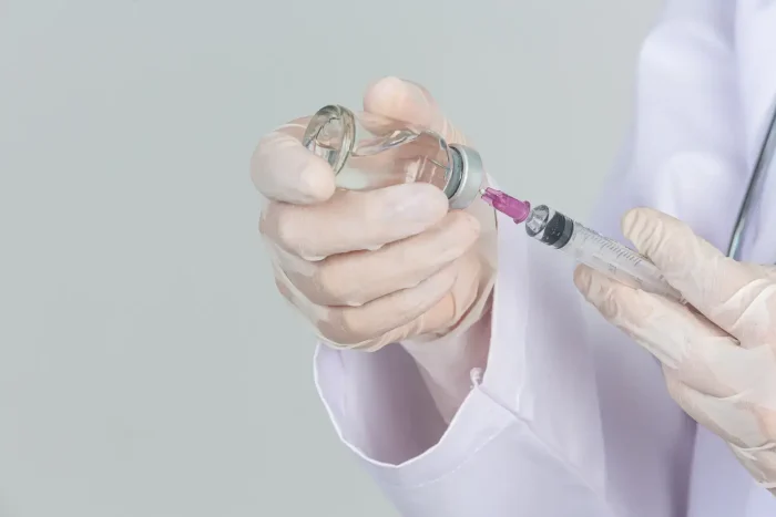 A person wearing gloves and a white lab coat holds a syringe and a vial. The syringe is being filled with liquid from the vial, suggesting preparation for an injection.