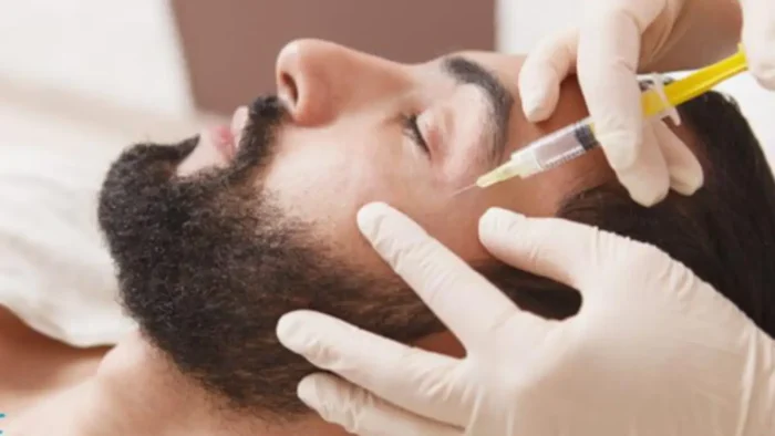 A person with a beard is lying on a flat surface, receiving an injection in the forehead area. The individual administering the injection is wearing white gloves and using a syringe.