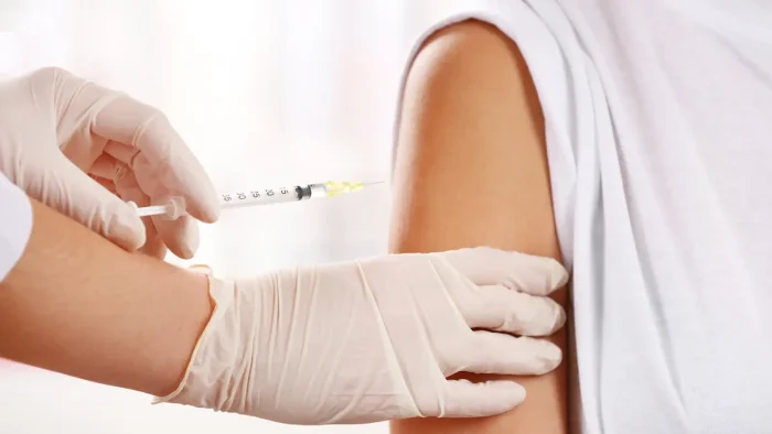 A healthcare professional wearing gloves administers a vaccine using a syringe into a person's upper arm. The person is wearing a white shirt, and only their arm is visible in the image.