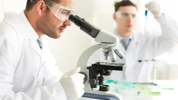 Two scientists in lab coats and safety goggles work in a laboratory. One is observing through a microscope, while the other is handling a pipette. Various lab equipment and glassware are visible on the table.