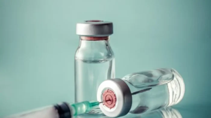 Two transparent vials with silver caps, one upright and one on its side, are placed on a light blue surface. A syringe with a green needle is positioned in front, partially out of focus.