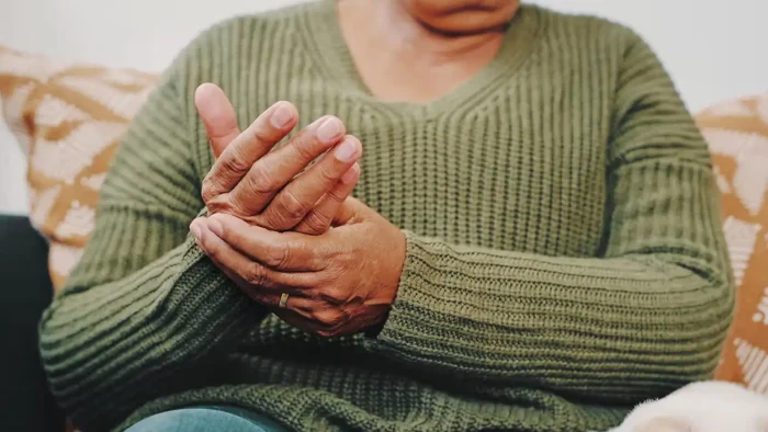 An individual wearing a green knit sweater is seated, holding their right hand with their left. The image focuses on the hands, suggesting care or discomfort. The background includes a pillow with a subtle pattern.