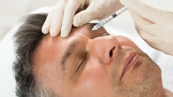 Person receiving an injection on the forehead by a gloved professional, lying down with eyes closed.