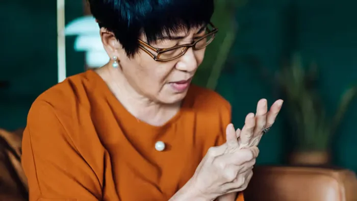 Elderly woman in glasses and an orange shirt looks down, holding and massaging her hand with a pained expression, suggesting discomfort or arthritis, against a blurred indoor background.