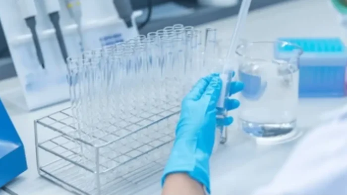 A person wearing blue gloves is using a pipette with a set of empty test tubes in a rack. Nearby, a beaker filled with liquid is on the lab bench. The scene depicts a laboratory setting.