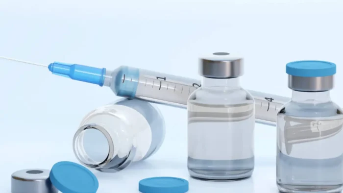 Vials with blue caps and a syringe with a needle lie on a white surface. The syringe is filled with clear liquid, and one of the vials is tipped over, with its cap detached. The background is a soft blue.