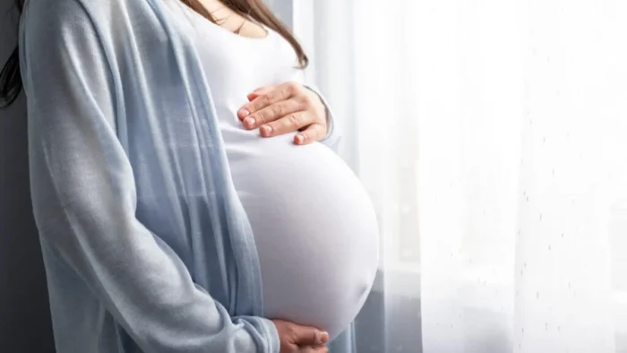 A pregnant woman gently cradles her belly while standing by a window. She is wearing a light cardigan over a white top, with soft daylight streaming in through white curtains.