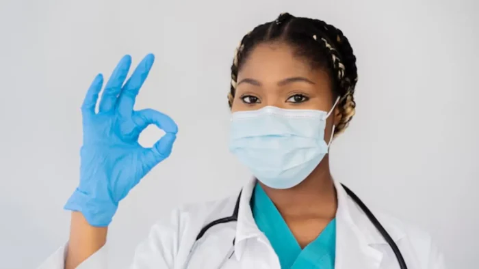 A healthcare professional wearing a surgical mask, blue gloves, teal scrubs, and a white coat is making an "OK" gesture with their right hand. The background is plain and light-colored.
