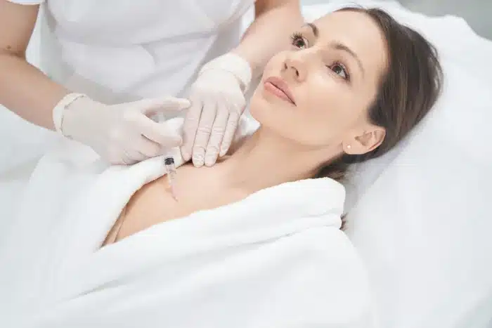A woman in a white robe is lying down while a medical professional wearing gloves performs an injection on her upper chest area. The woman looks relaxed as she receives the treatment.