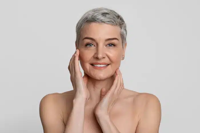 Smiling person with short gray hair touching their face, posing against a plain light background. They have a confident and positive expression.