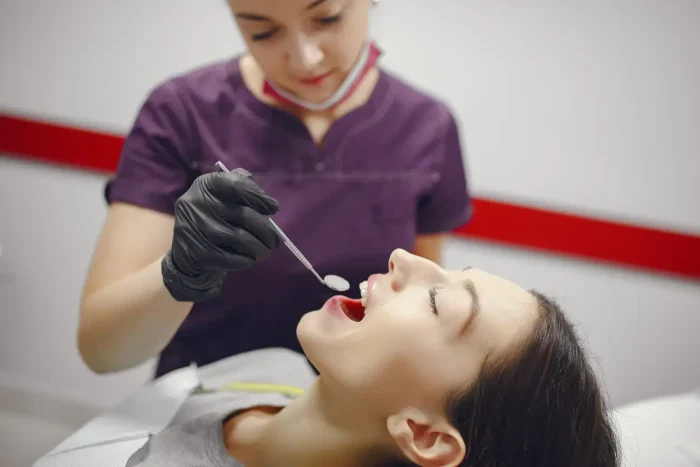 A dental professional in a deep purple uniform examines a patient's teeth using a dental mirror. The patient is reclining with their mouth open in a dental chair. The professional is wearing gloves and a protective face mask.