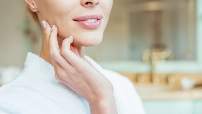 Close-up of a person wearing a white robe, touching their cheek with one hand. They have light skin, pink lipstick, and short nails. The background is a blurred bathroom setting, creating a serene and relaxed atmosphere.