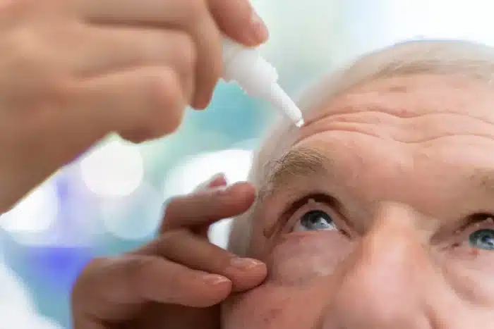 Close-up of an elderly person receiving eye drops. A hand holds the bottle above their open eye, while the other hand gently pulls down the lower eyelid. The background is softly blurred.
