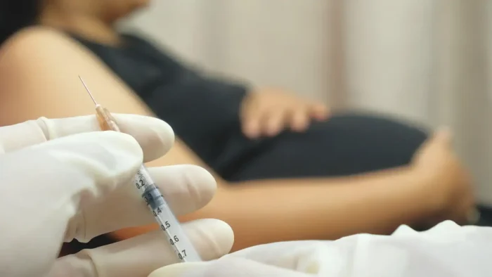 Close-up of a gloved hand holding a syringe in the foreground, with a pregnant person in a dark top sitting in the background, one hand resting on their belly. The setting appears clinical or medical.