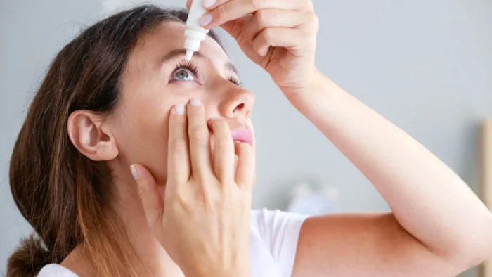 A person with brown hair uses an eye dropper to apply eye drops. They are looking upward and gently pulling down their lower eyelid. The background is softly blurred, and the person is wearing a white top.