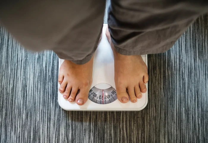 Person wearing gray pants standing on a white analog bathroom scale. The dial is visible, showing the weight measurement. The background is a textured, gray carpeted floor.
