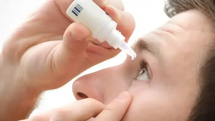 A person applying eye drops to their right eye. The close-up image shows hands holding a small bottle with the applicator tip near the eye, squeezing a drop of liquid towards the open eye. The background is a soft white.