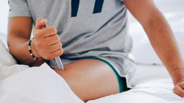 Person sitting on a bed, administering an injection into their thigh. They are wearing a gray shirt and green shorts. The setting appears casual and relaxed, with the focus on the act of injecting.