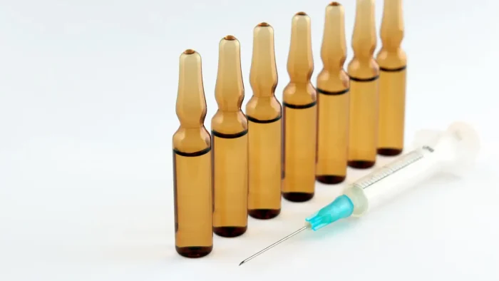 Six amber glass ampoules are lined up in a row next to a medical syringe with a blue-tipped plunger on a plain white background.