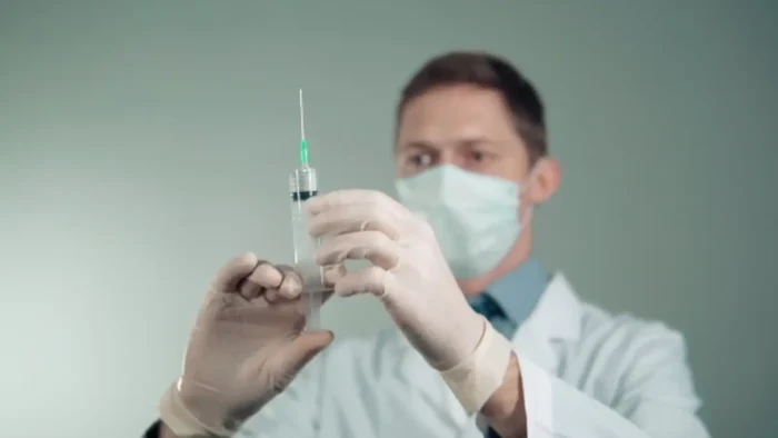 A person wearing a white lab coat, face mask, and gloves is holding a syringe, carefully examining it against a neutral background. The focus is on the syringe, with the individual blurred in the background.