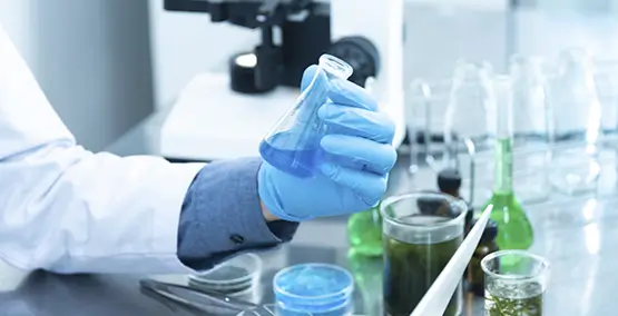 A scientist wearing a white lab coat and blue gloves holds a flask containing a blue liquid. Various lab equipment, including glass containers with different colored liquids and a microscope, are visible on the shiny metal table.