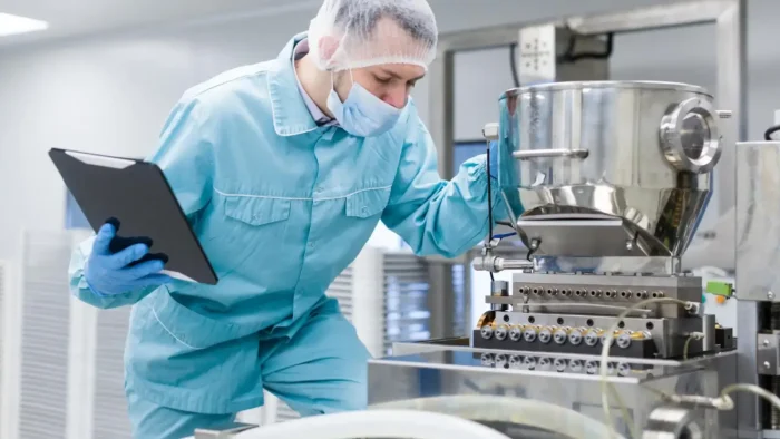 A technician in protective clothing, face mask, and hairnet inspects a stainless steel machine in a laboratory or industrial setting, holding a clipboard. The environment is clean and sterile, suggesting a focus on safety and precision.
