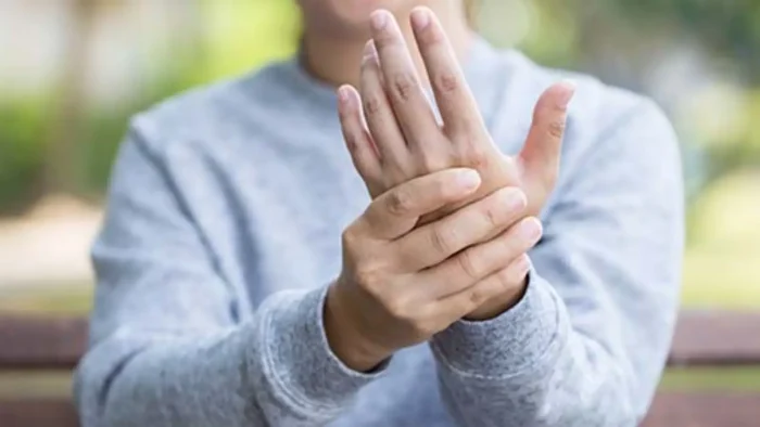 A person wearing a gray sweatshirt holds their right hand with their left, as if massaging it, while sitting outdoors on a bench. The background is blurred with green foliage.