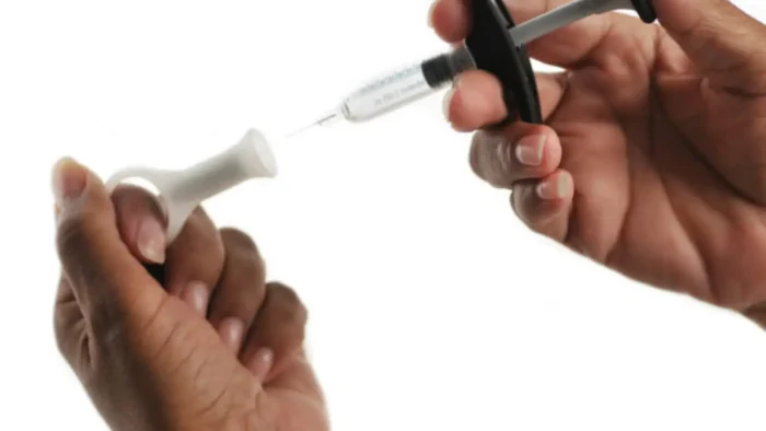 Two hands are shown: one holding a syringe filled with liquid, and the other gripping a device that appears to be a nasal spray. The background is plain white.