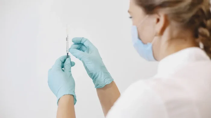 A healthcare worker wearing a mask and blue gloves is holding a syringe, presumably preparing for vaccination. The background is plain and white, giving a clinical atmosphere.