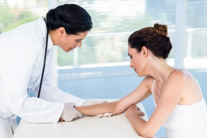 A doctor wearing a white coat and gloves examines a patient's arm. The patient, sitting on an examination table, looks at the doctor's hands. The background is a bright room with blurred windows.