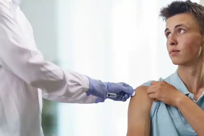 A young man looks to the side while receiving a syringe injection in his upper arm from a healthcare professional. The professional wears a white coat and blue gloves, pressing a swab on the man's arm. The background is softly blurred.