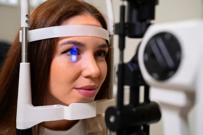 A woman undergoes an eye exam using a slit lamp. Her face is framed by the lamp's head support, and a blue light is focused on her eye. She appears attentive and the background is softly blurred.