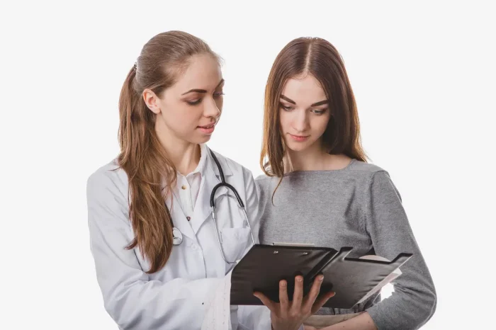 A medical professional in a white coat and stethoscope showing documents on a clipboard to a woman with long brown hair wearing a gray top. Both are focused on the paperwork against a white background.