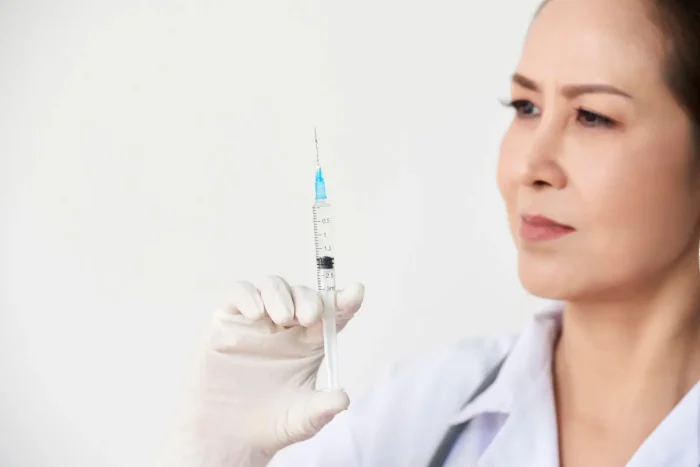 A person wearing a white coat and gloves is holding up a syringe and looking at it intently. The background is plain white.