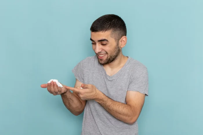 A man with short dark hair and a beard is smiling while holding a pile of shaving cream in his right hand and pointing at it with his left index finger. He is wearing a gray T-shirt against a light blue background.