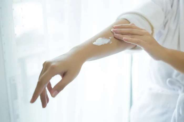 A person applying white lotion to their forearm in front of a softly lit window. They are wearing a white robe, and their fingers gently spread the cream over the skin. The background is bright and blurred.