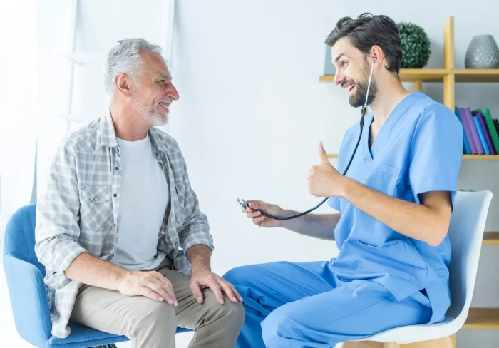 An older man sits and smiles at a healthcare professional in blue scrubs, who is holding a stethoscope and giving a thumbs up. They appear to be having a friendly, positive conversation in a bright room.