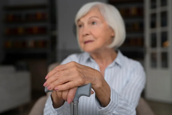 An elderly woman with short white hair, wearing a striped shirt, sits indoors holding a cane and looks thoughtfully into the distance. Her hands are resting on top of the cane.