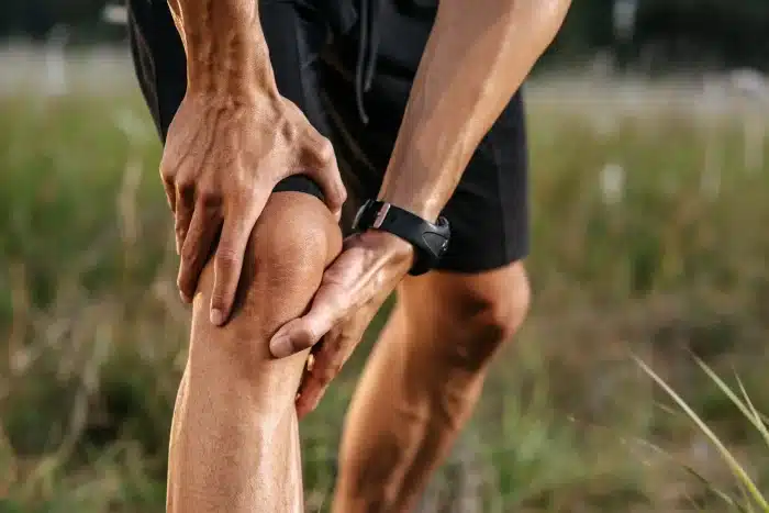 A person wearing black shorts holds their knee with both hands, appearing to be in pain, while standing outdoors on grass.