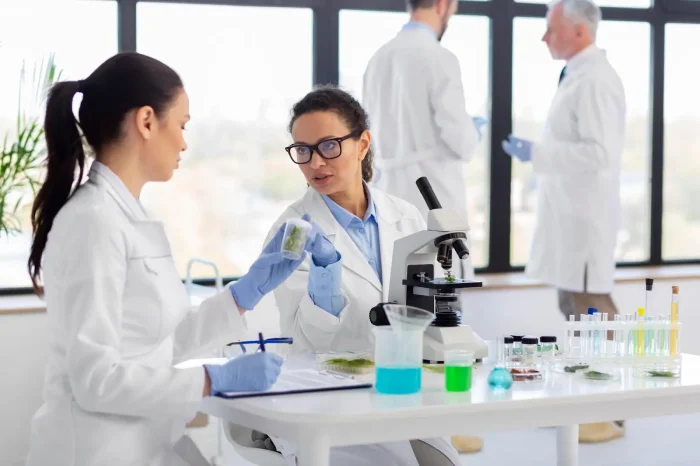 Two scientists in lab coats and gloves discuss a sample next to a microscope. A table with test tubes and beakers is in the foreground. In the background, two other scientists converse near a window.