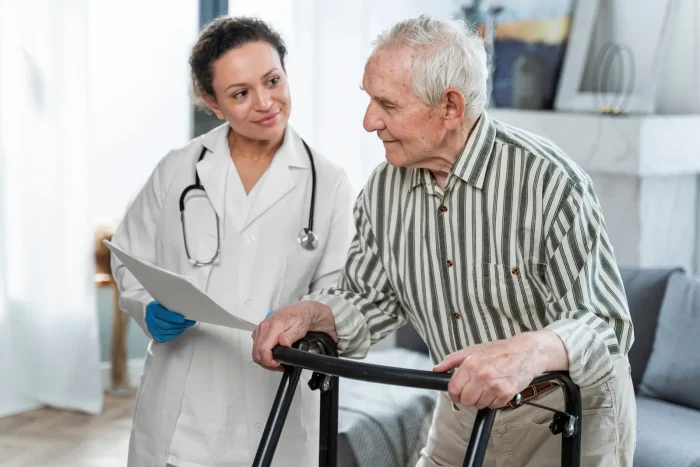A doctor with a stethoscope and medical gloves talks to an elderly man using a walker. The doctor is holding a clipboard and smiling at the man in a home-like setting.