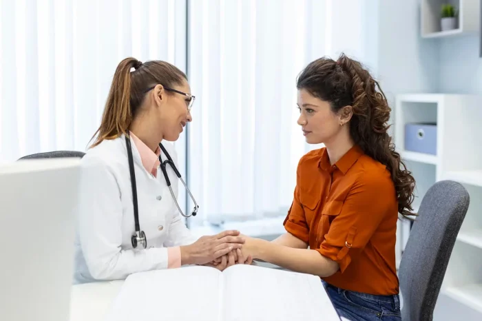 A doctor in a white coat is sitting at a desk, holding hands with a woman in an orange blouse, providing comfort and support. The office has a bright and clean appearance with a large window in the background.
