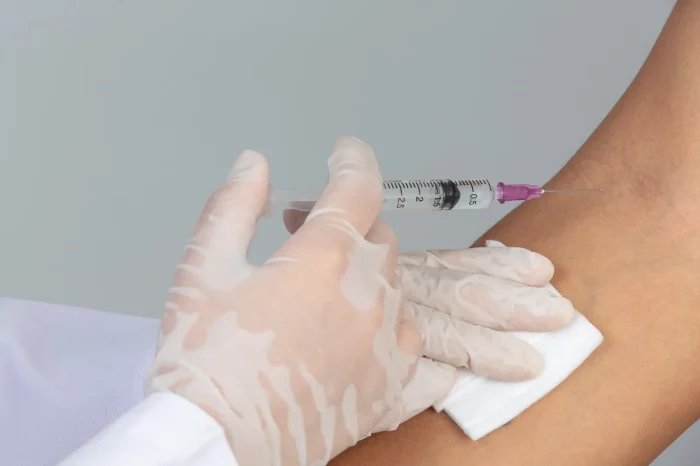 A medical professional wearing white gloves prepares to inject a syringe into a person's arm, pressing a cotton pad against the skin.