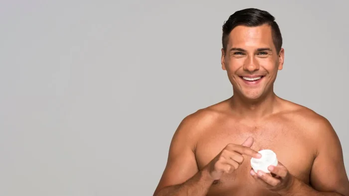 A shirtless man smiling while holding a white container and applying cream to his finger. The background is plain gray.