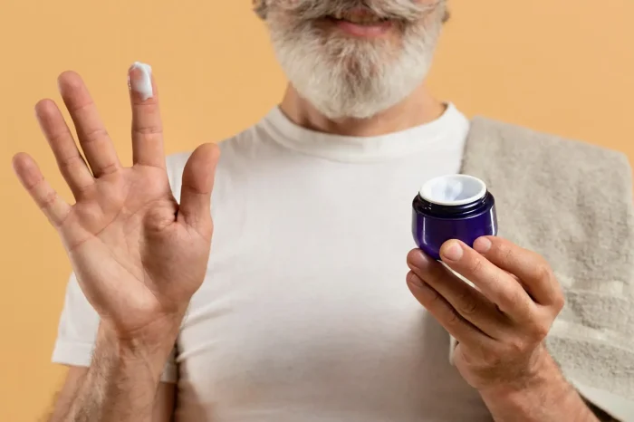 A smiling person with a beard holds an open jar of cream in one hand and has a dab of cream on the finger of the other hand. They are wearing a white shirt and have a towel draped over their shoulder. The background is a warm beige color.